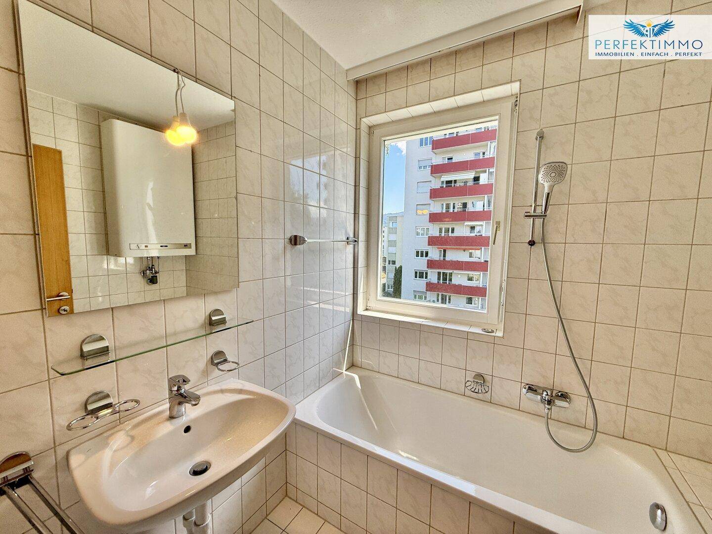 Gefliestes Badezimmer mit Badewanne, Waschbecken, Spiegel und Fenster für Tageslicht.
