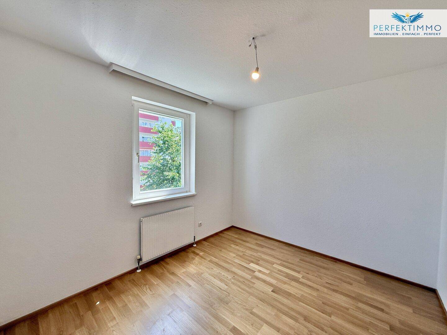 Helles Schlafzimmer mit Parkettboden, weiß gestrichenen Wänden und Fenster.