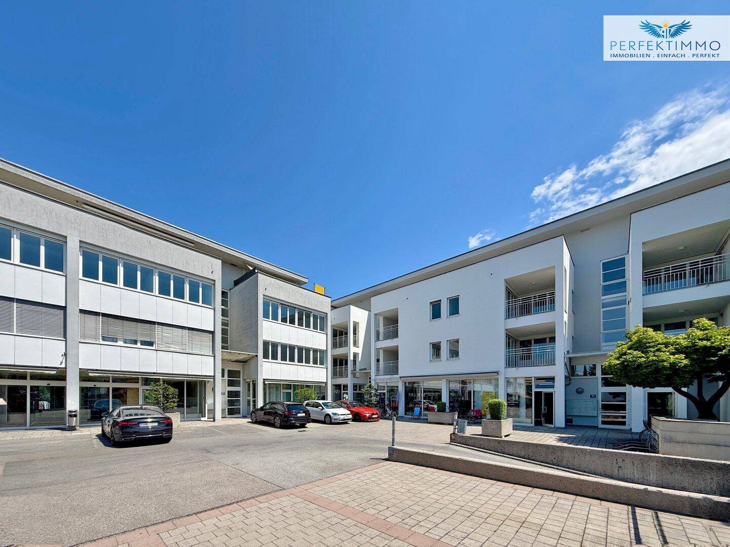 Moderner Gebäudekomplex mit Geschäften im Erdgeschoss und Wohnungen darüber, umgeben von Parkplätzen.