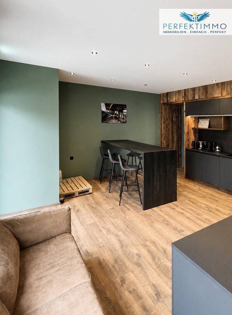 Offener Bürobereich mit Stehtisch, Barhockern und einer bequemen Couch vor grüner Wand.