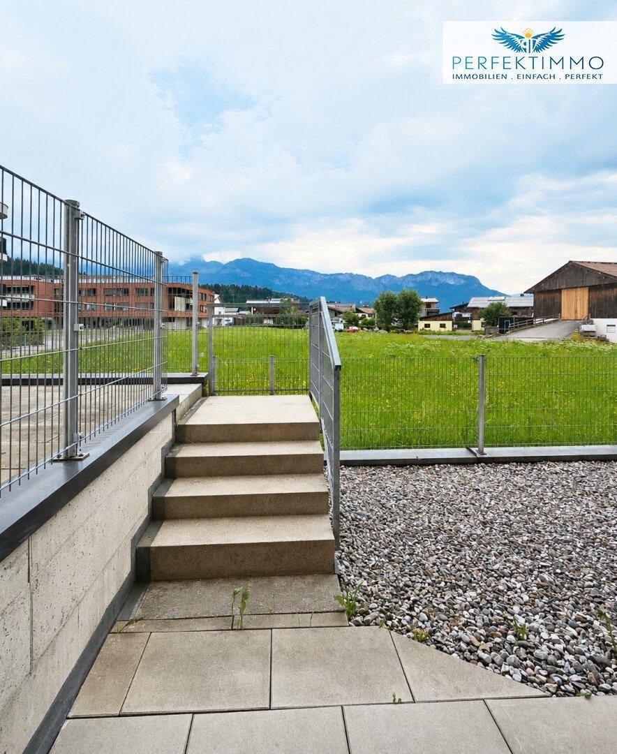 Außenbereich mit Treppe, Kiesfläche und Blick auf eine grüne Wiese und Berge.