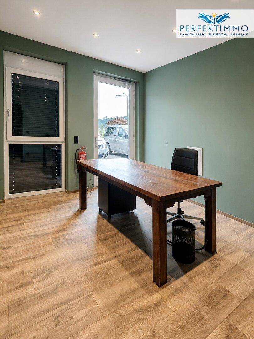 Helles Büro mit Holztisch, Bürostuhl und Fenster mit Außenjalousien.