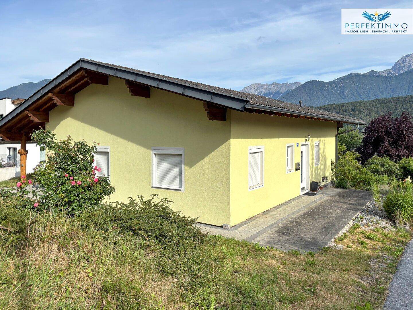 Seitenansicht des Hauses mit gelber Fassade und gepflastertem Zugang, umgeben von Bergen.