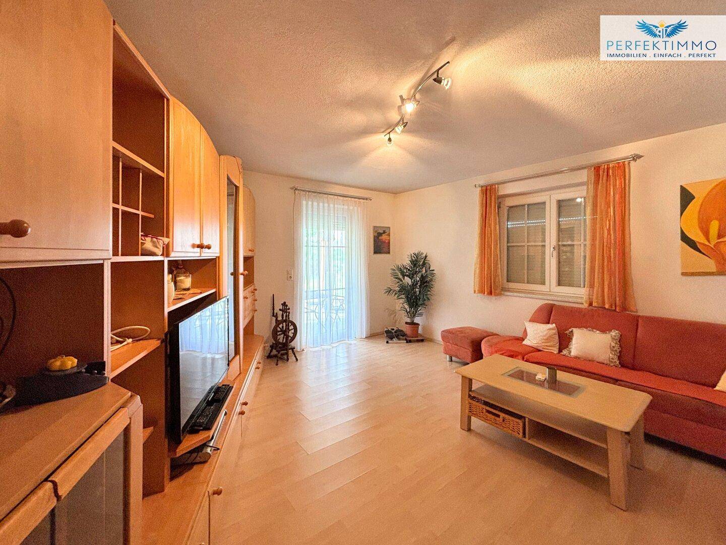 Gemütliches Wohnzimmer mit hellem Holzboden, rotem Sofa und Zugang zum Außenbereich.