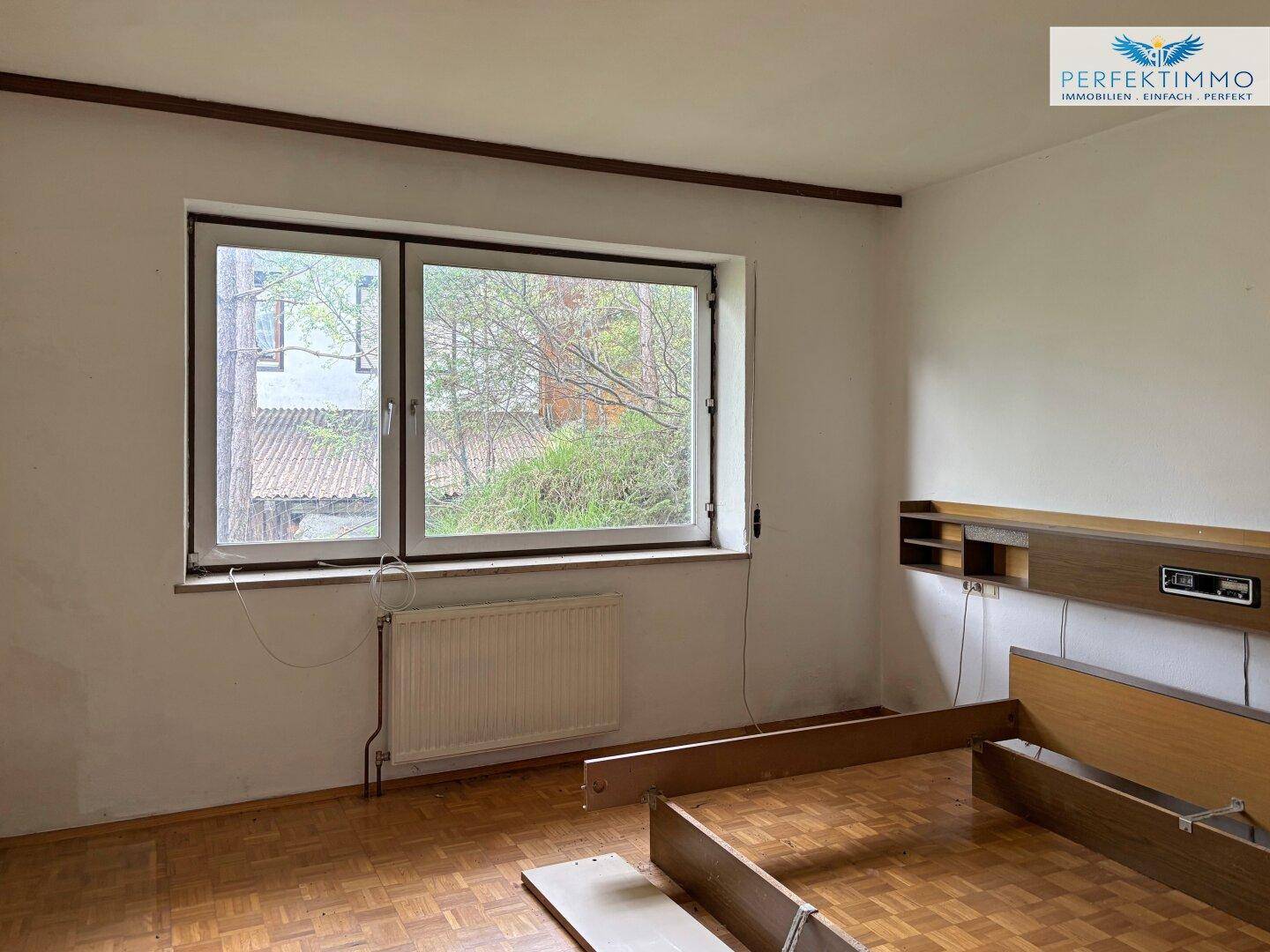 Unmöbliertes Schlafzimmer mit Parkettboden und Fenster, das einen Blick ins Grüne bietet.