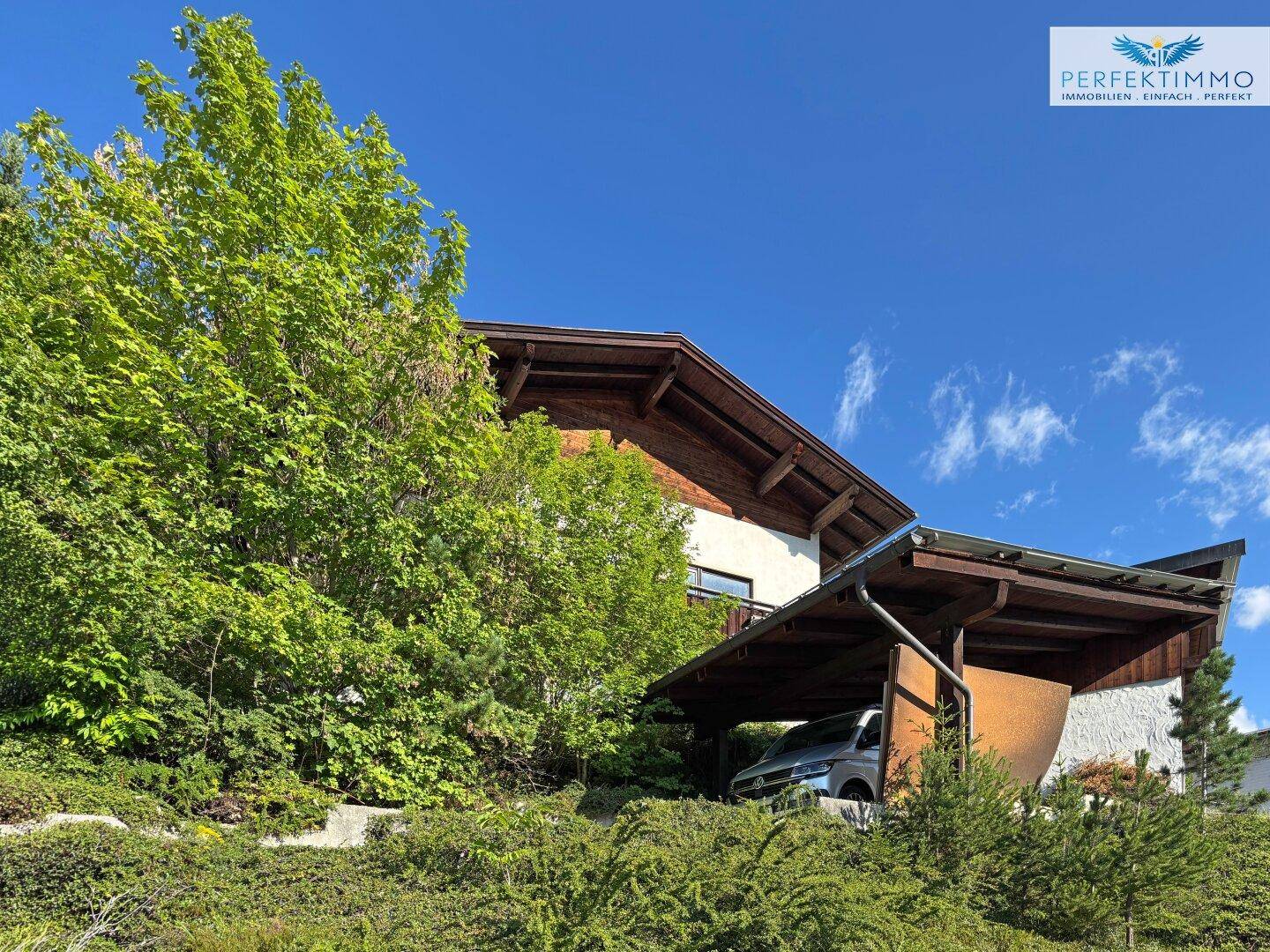 Ansicht des Hauses mit Carport und üppiger grüner Umgebung unter blauem Himmel.