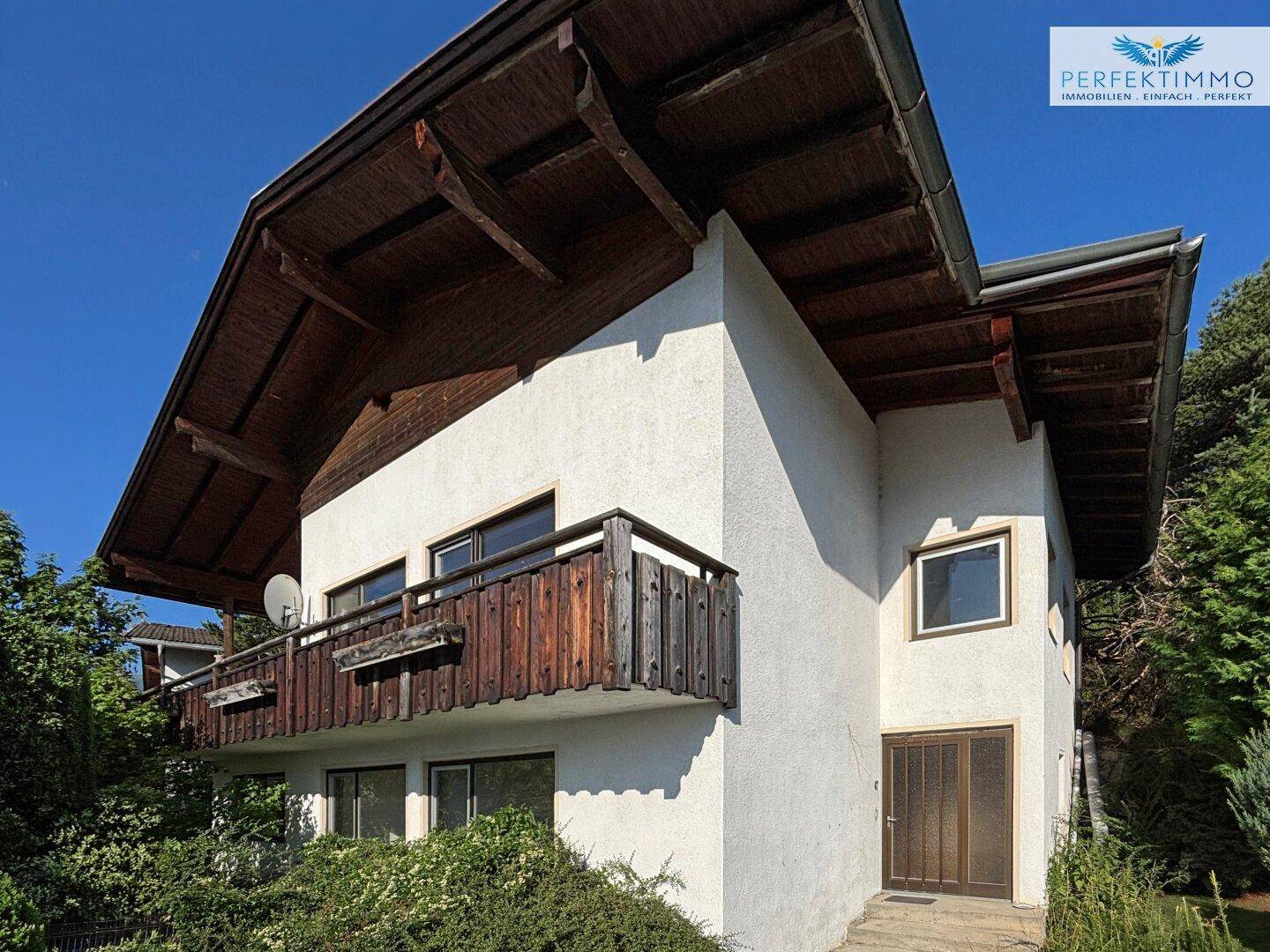 Hausfassade mit Holzbalkon und traditionellem Satteldach, umgeben von grüner Bepflanzung.