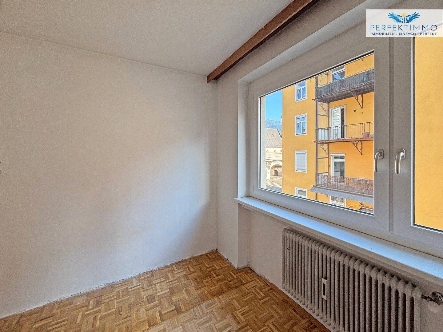 Heller, leerer Raum mit Parkettboden und einem Fenster mit Blick auf die Stadt.