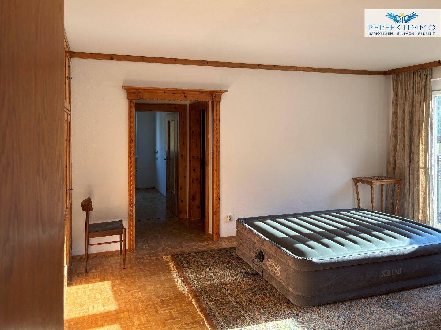 Schlafzimmer mit Parkettboden, Holztürrahmen und Zugang zu einem Balkon.