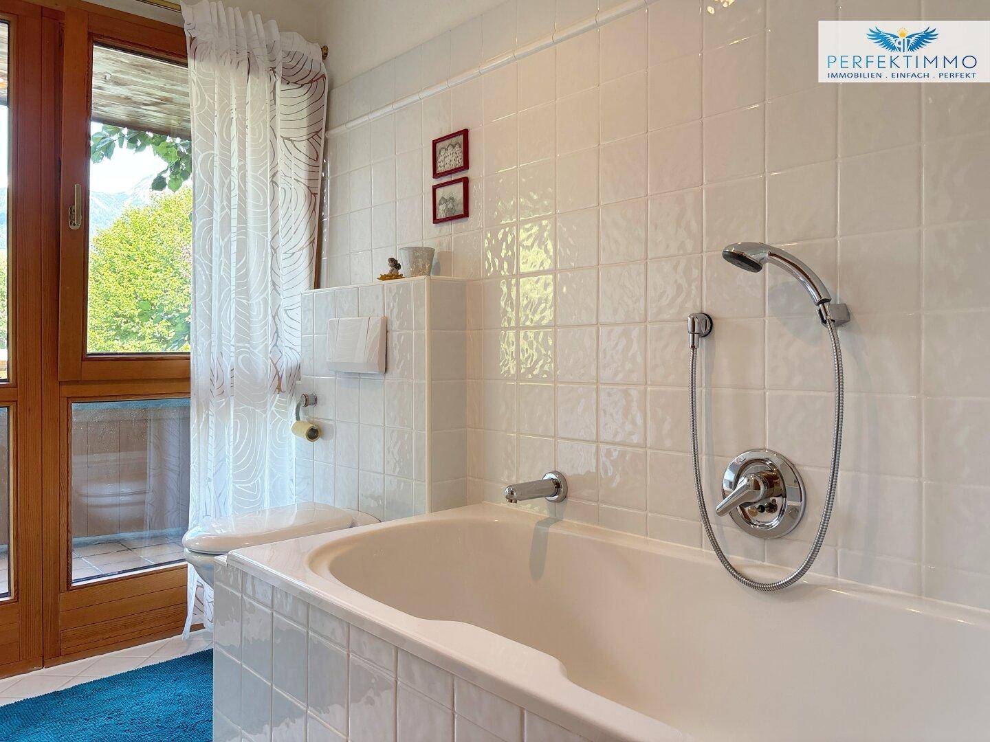 Badezimmer mit Badewanne, weißen Fliesen und Blick ins Grüne durch die Fenstertür.