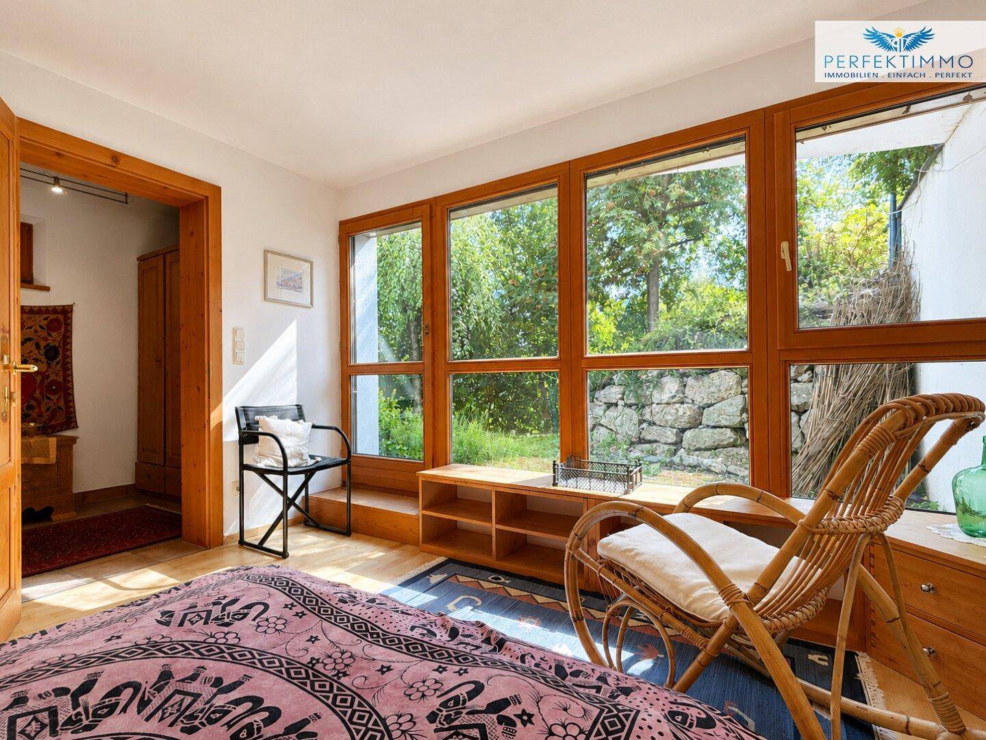 Helles Schlafzimmer mit großen Fenstern, Blick ins Grüne und Rattansessel.