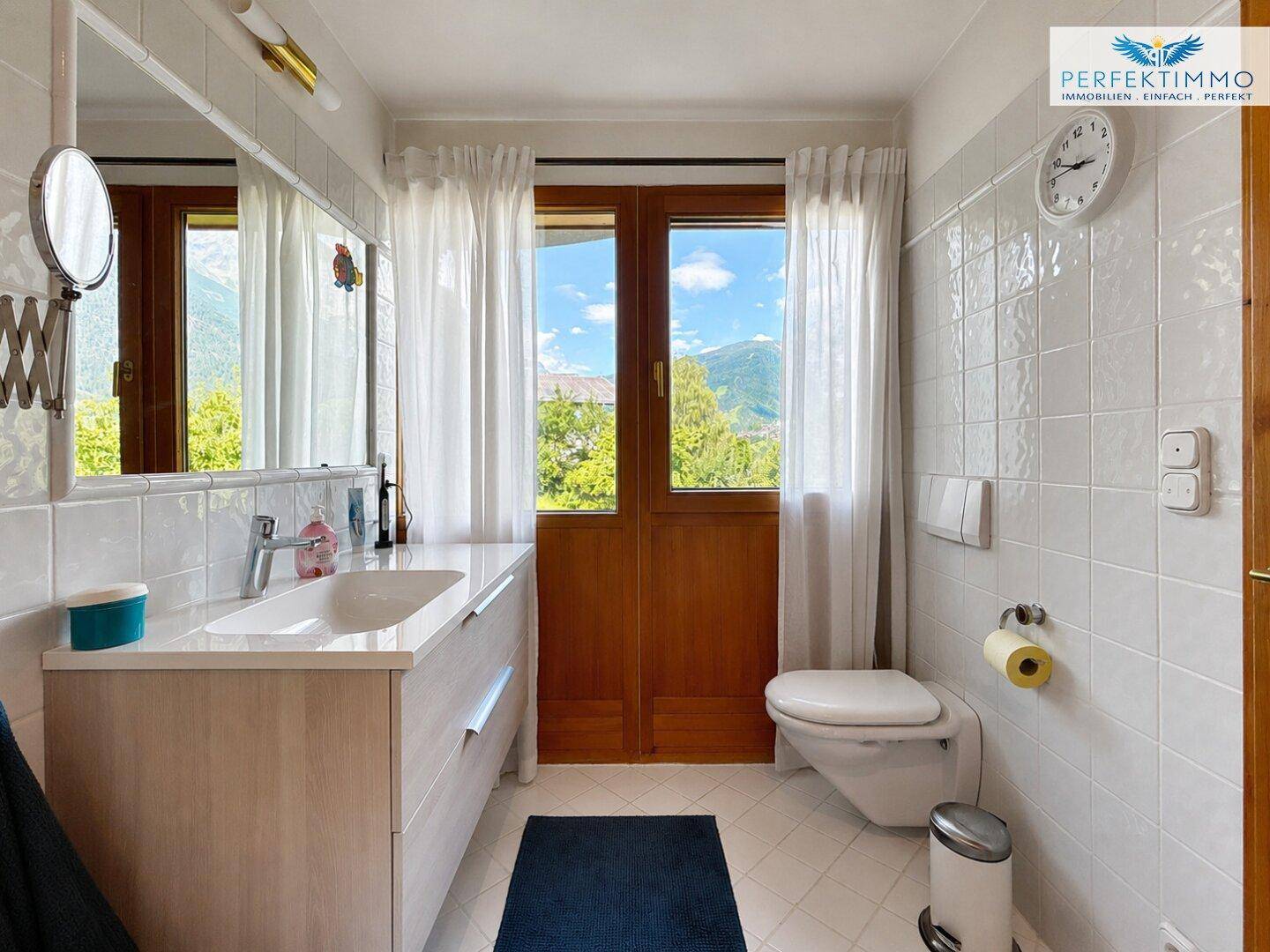 Helles Badezimmer mit Fenster, Waschtisch, WC und weißen Fliesen, bietet Bergblick.