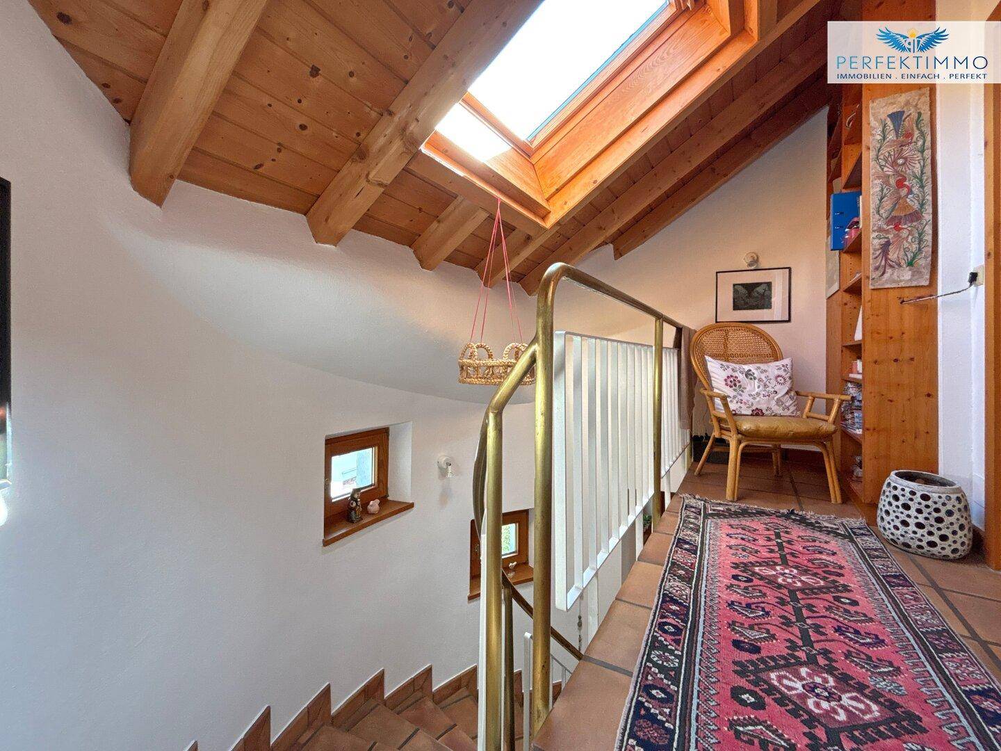 Heller Flur mit Holzbalkendecke, Dachfenster und Teppichläufer, der zu den oberen Etagen führt.