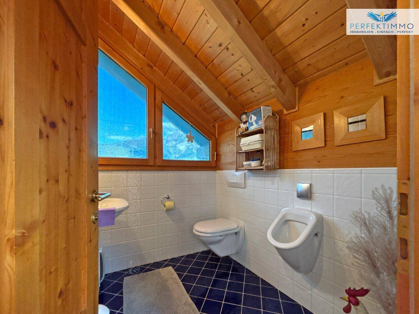 Kleines Gäste-WC mit Urinal, WC und Waschbecken, blauen Fliesen und Holzbalkendecke.