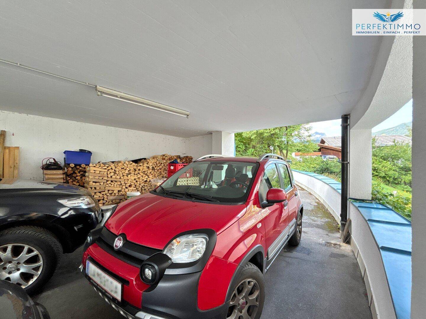 Überdachter Parkplatz für Fahrzeuge mit Holzstapel im Hintergrund und Blick ins Grüne.