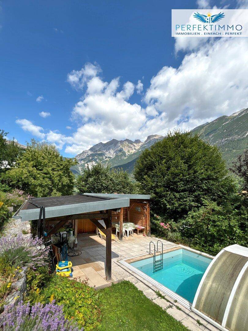 Gartenbereich mit Pool, überdachtem Sitzplatz und blühenden Pflanzen unter blauem Himmel.