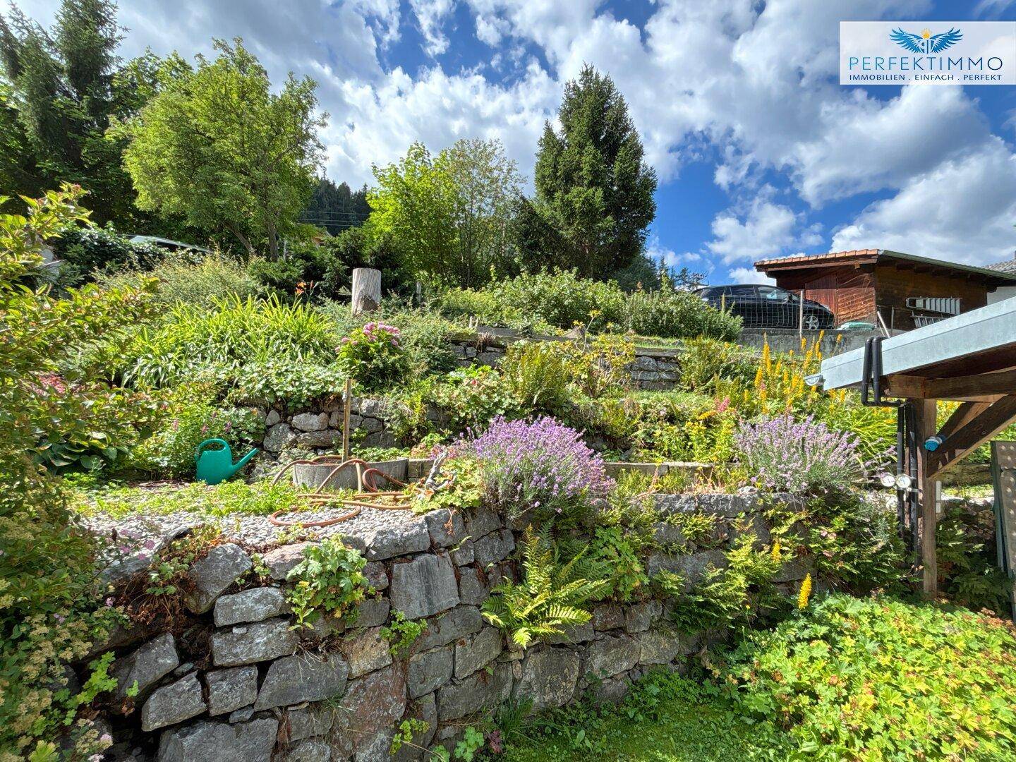 Terrassierter Gartenbereich mit Steinmauern, vielfältiger Bepflanzung und Blick auf die Umgebung.