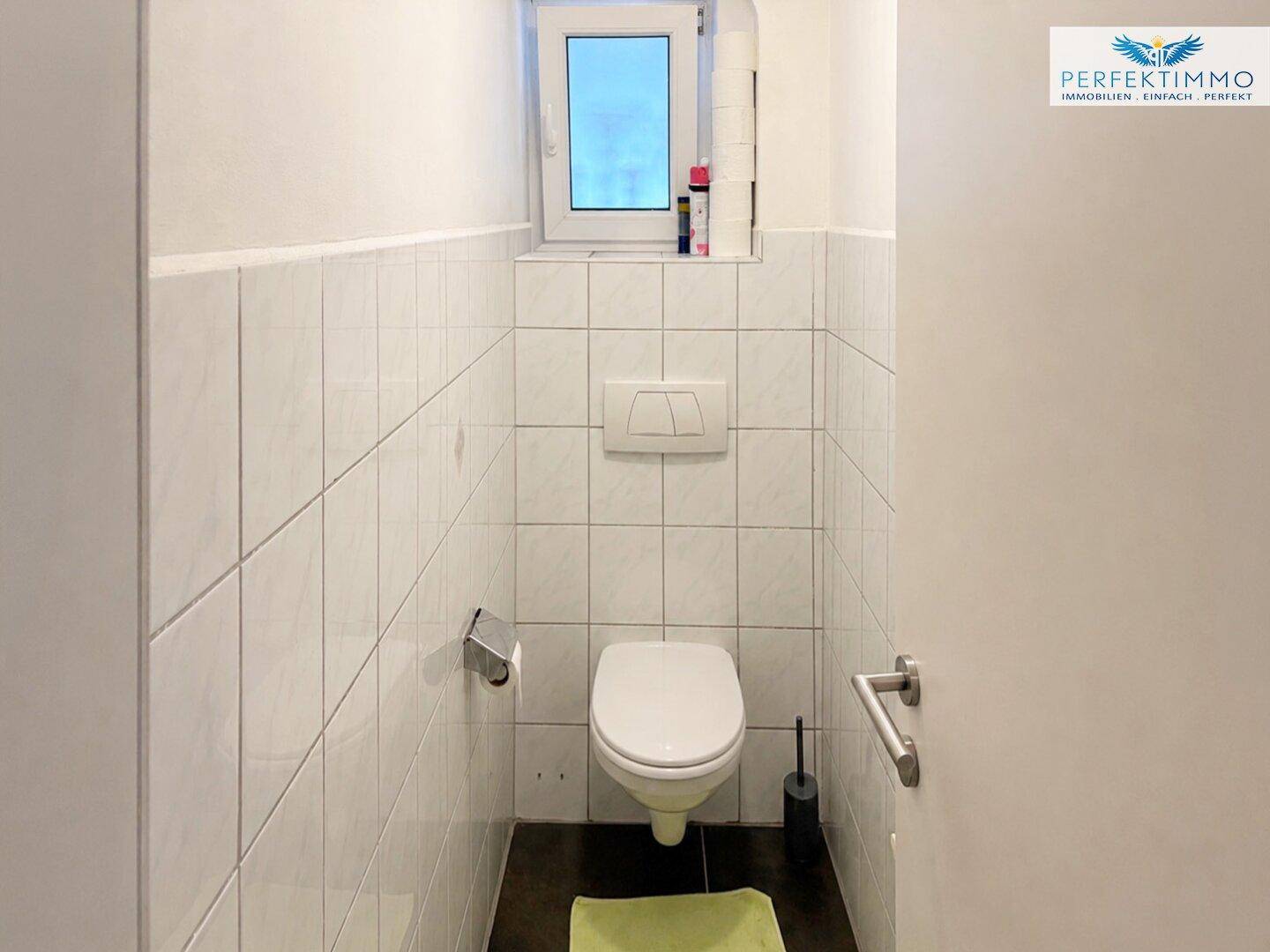 Separate Toilette mit Wand-WC und kleinem Fenster, weiß gefliest.