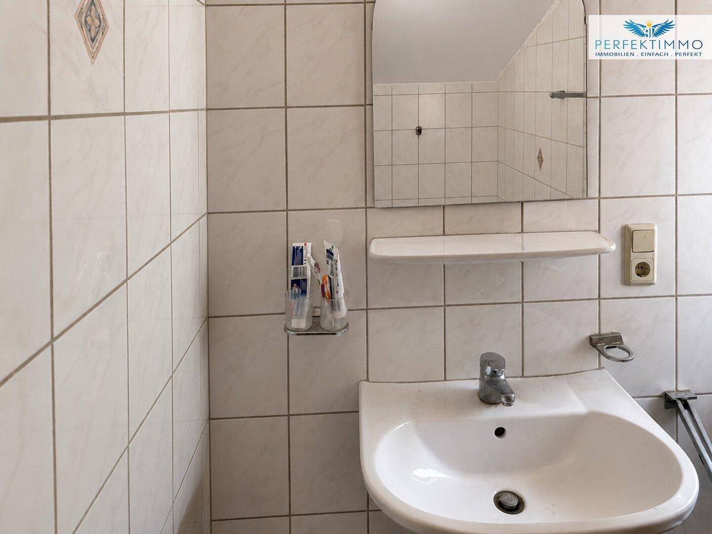 Helles Badezimmer mit Fliesenwänden, Waschbecken und Spiegel, ausgestattet mit Standardarmaturen.