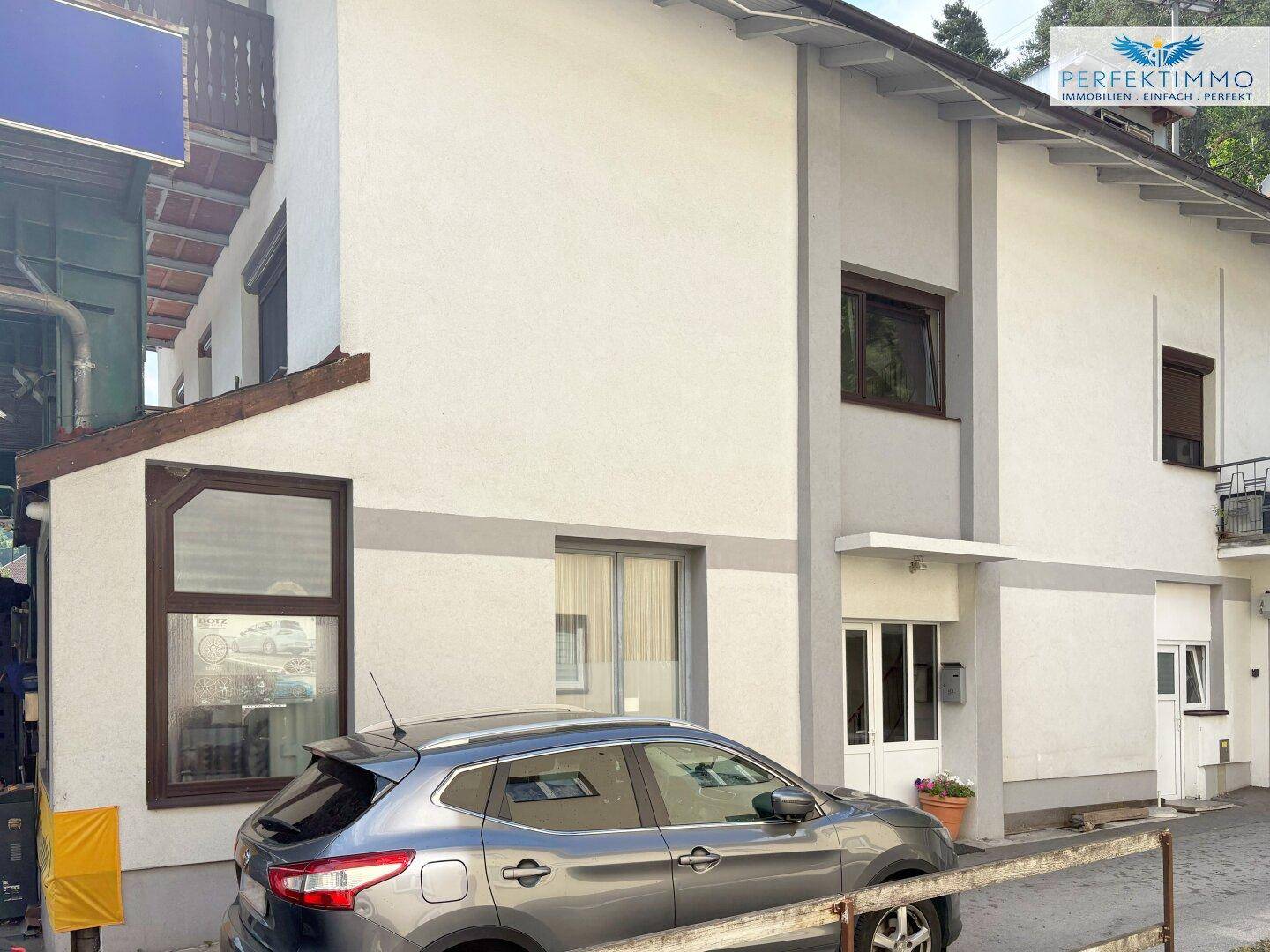 Fassade des Gebäudes mit geparktem Auto und modernen Fenstern, umgeben von Grün.