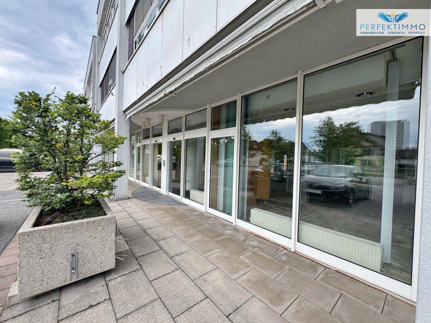 Ansicht der Geschäftsfassade mit großen Glasfronten und einem gepflegten Blumenkübel am Eingangsbereich.