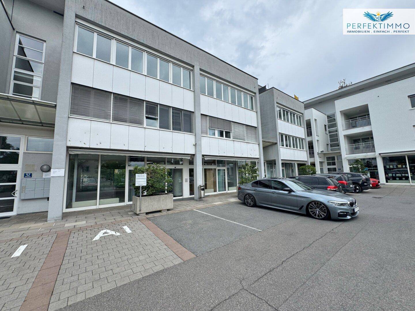 Außenansicht eines modernen Geschäftsgebäudes mit geparkten Autos und großen Fenstern, unter bewölktem Himmel.