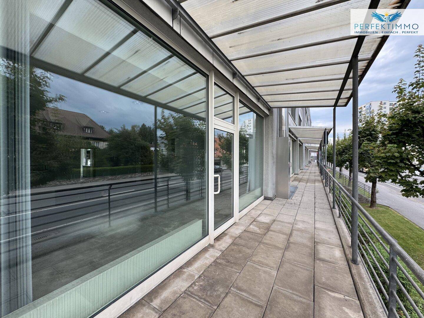 Überdachter Außenbereich entlang der Geschäftsfassade mit Blick auf die Straße und Bäume, bietet Schutz.