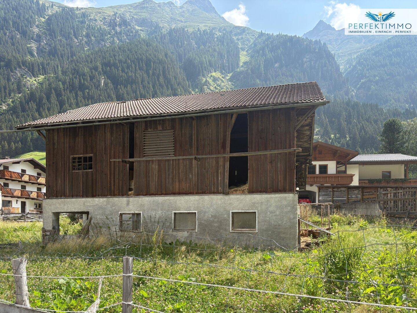 Detailansicht eines alten Holzgebäudes mit Steinfundament, eingebettet in eine grüne Berglandschaft.
