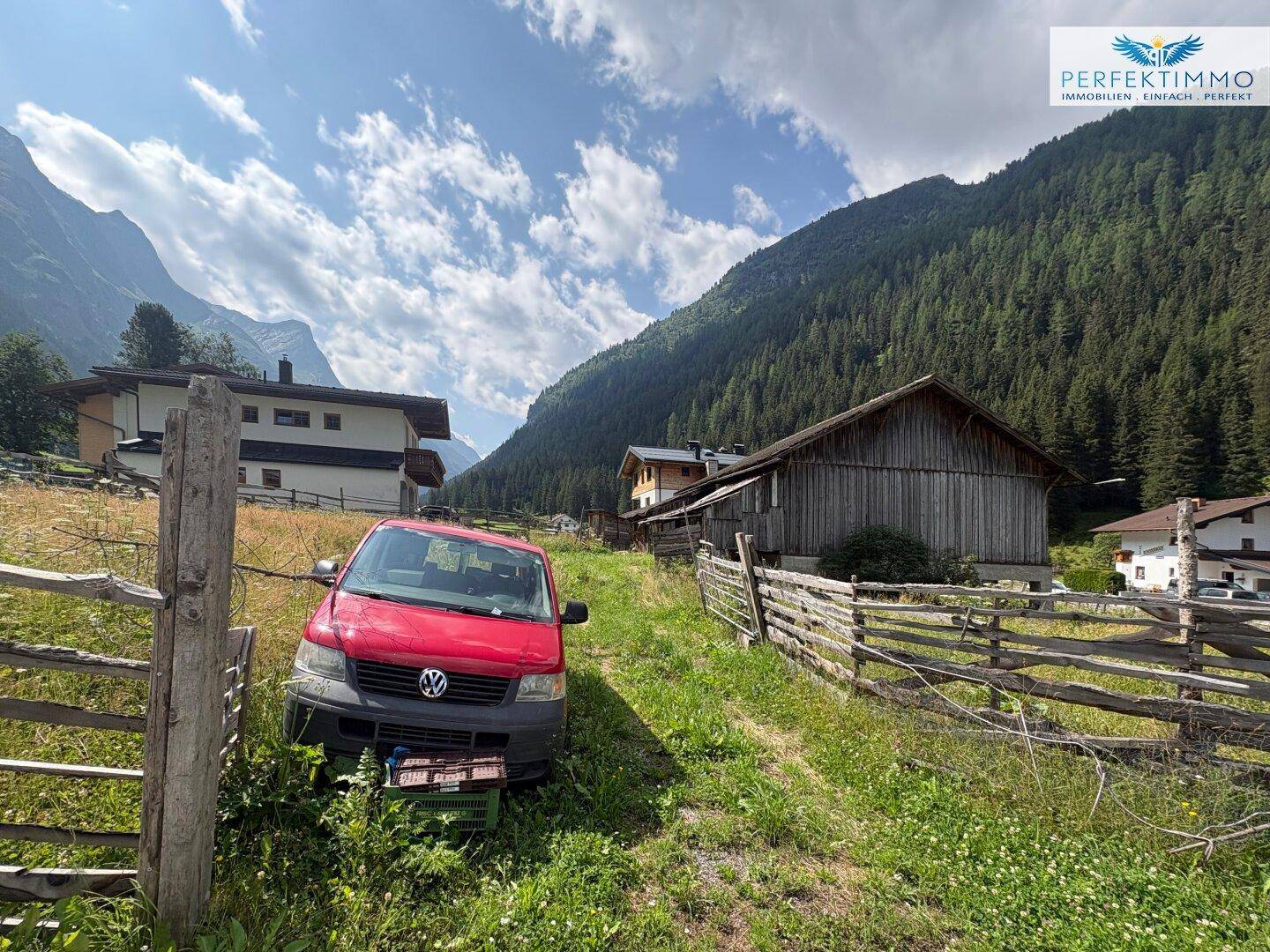 Roter Van parkt auf einer Wiese neben einem traditionellen Holzhaus mit Bergen im Hintergrund.