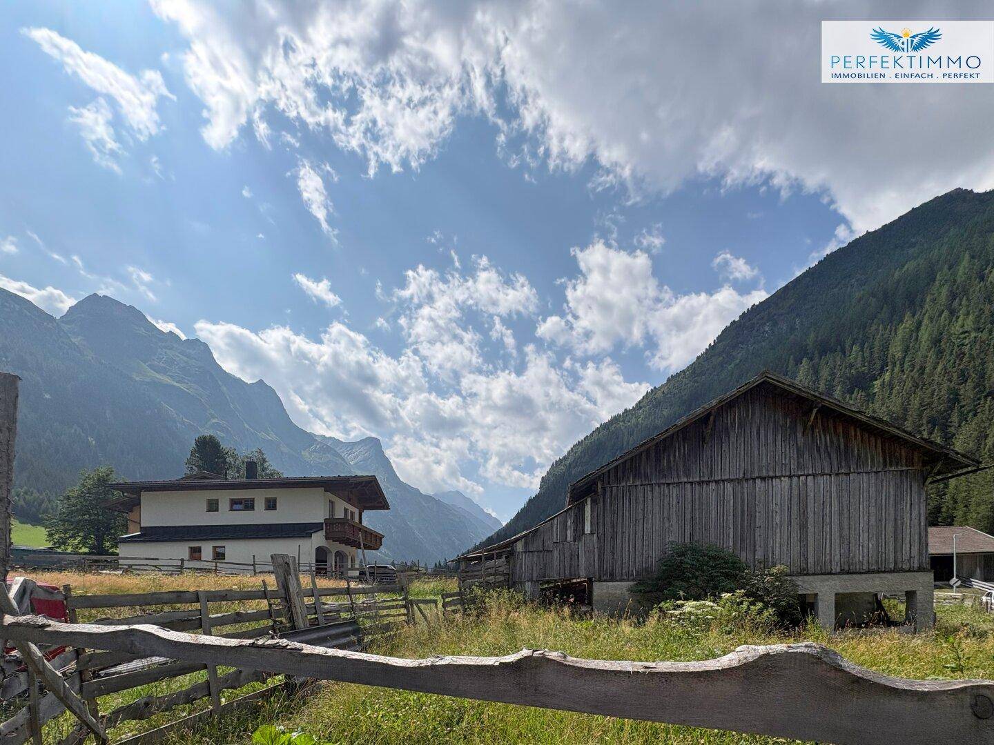 Weitläufige Ansicht eines Holzgebäudes und eines Wohnhauses in einem Tal mit hohen Bergen.