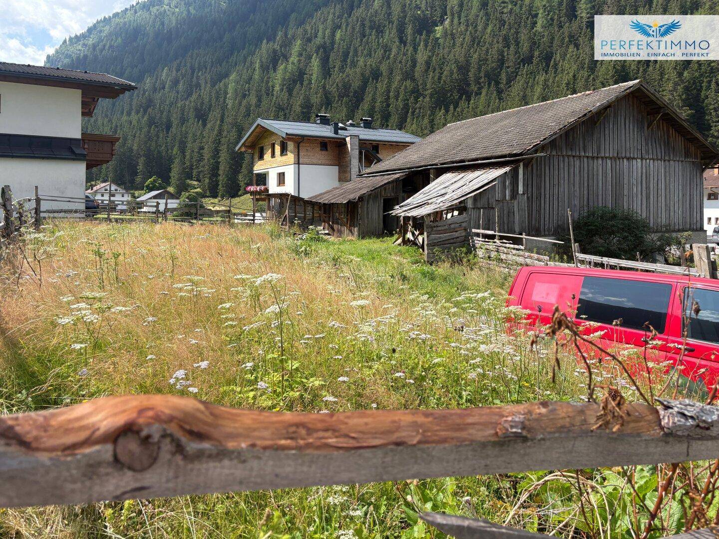 Ländliche Szene mit einem roten Fahrzeug auf einer blühenden Wiese vor traditionellen Gebäuden.