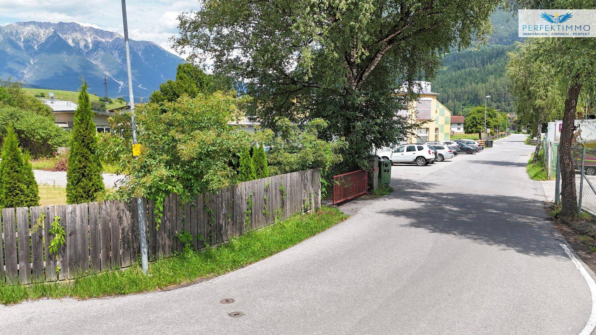 Blick auf eine Straße mit Holzzaun, Bäumen und einer beeindruckenden Bergkulisse im Hintergrund.