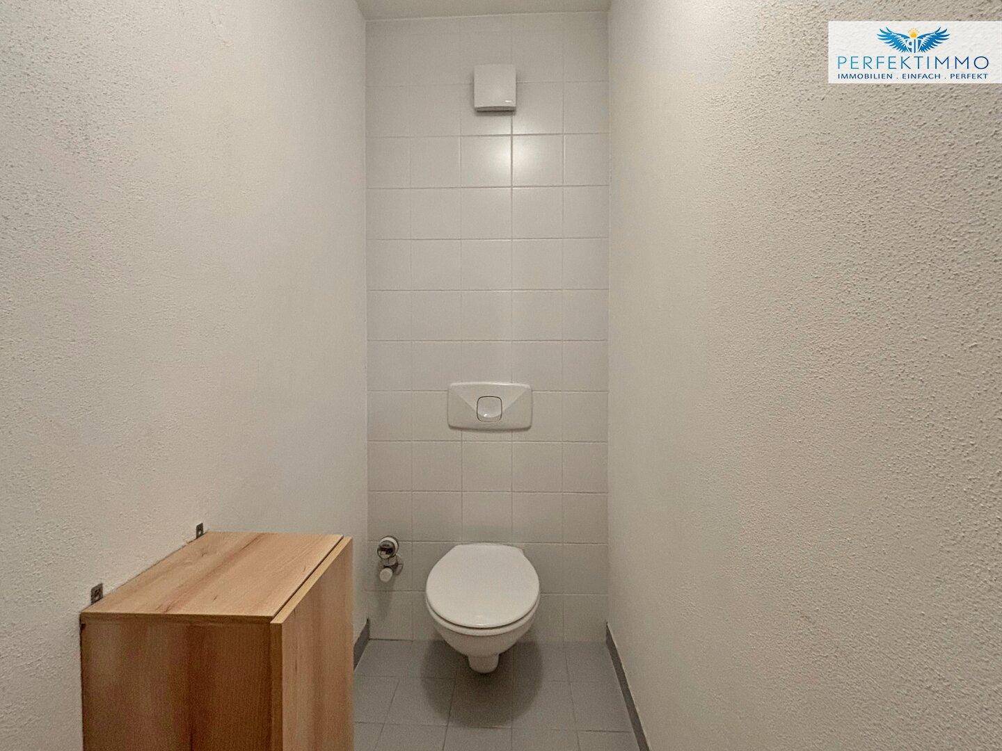 Separates WC mit weißer Toilette, Fliesenwand und kleinem Holzschrank.