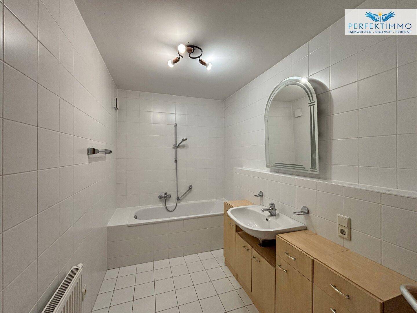 Helles Badezimmer mit weißen Fliesen, Badewanne mit Duschfunktion und Holzunterschrank.