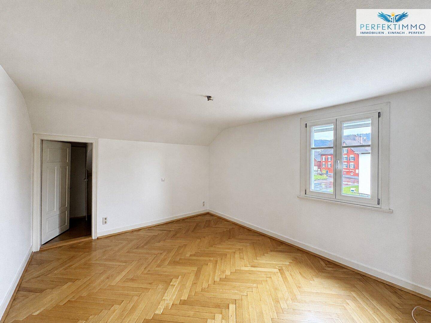 Helles Schlafzimmer mit Fischgrätparkett, Dachschräge und Fenster, das viel Tageslicht hereinlässt.