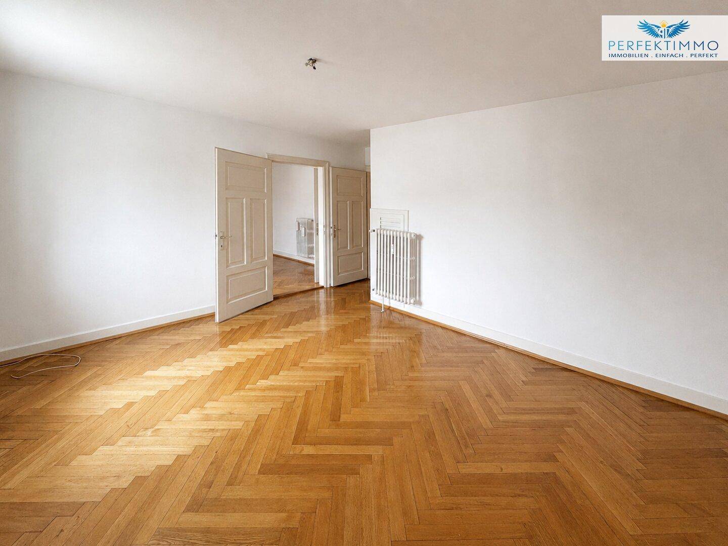 Geräumiges Wohnzimmer mit elegantem Fischgrätparkett und Zugang zu weiteren Räumen.