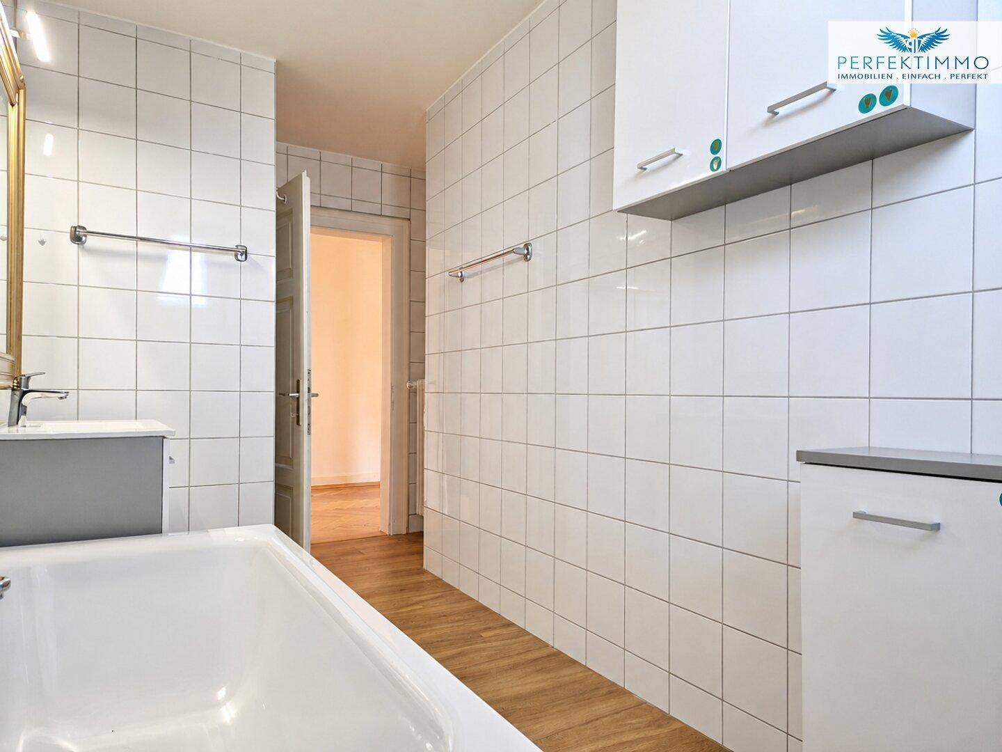 Helles Badezimmer mit weißen Fliesen, Badewanne und Blick auf den angrenzenden Raum.
