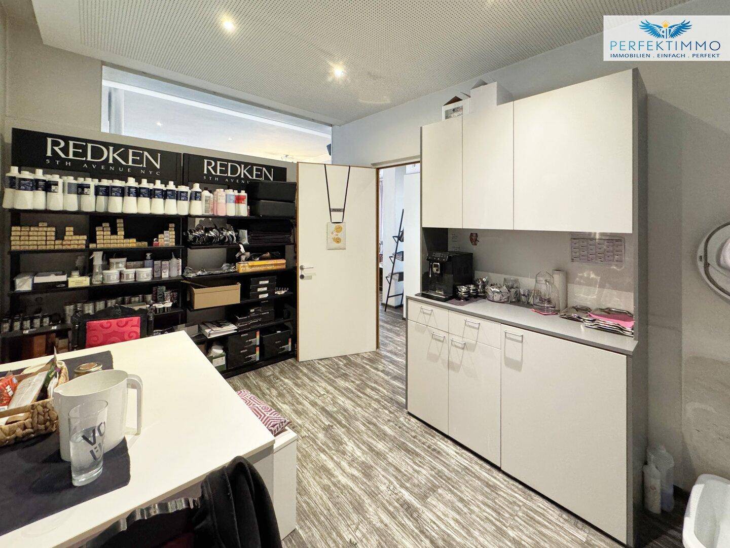 Kleine Küchenzeile mit weißen Schränken, Kaffeemaschine und Regalen für Salonprodukte und Utensilien.