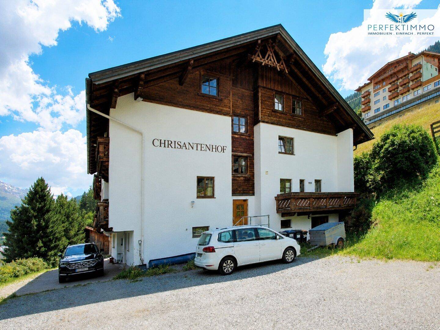 Alpines Gebäude mit Holzelementen und weißer Fassade, umgeben von grünen Hängen und Bergen.
