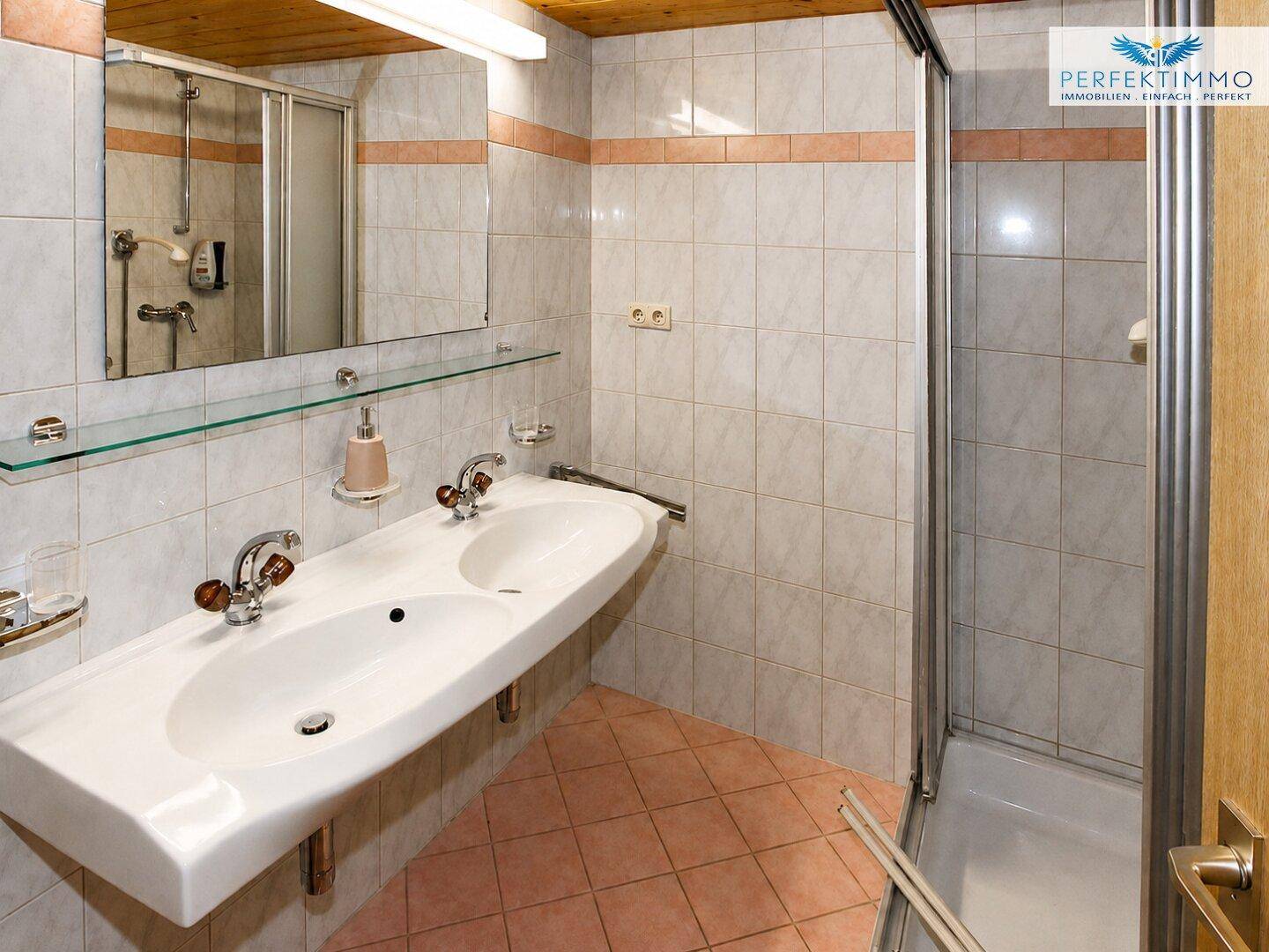 Helles Badezimmer mit Doppelwaschbecken, Duschkabine und hellen Fliesen.