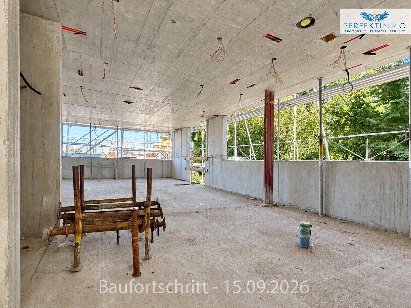 Offener Rohbau-Innenraum mit großen Fensteröffnungen, die einen Blick ins Grüne und auf das Baugerüst bieten.
