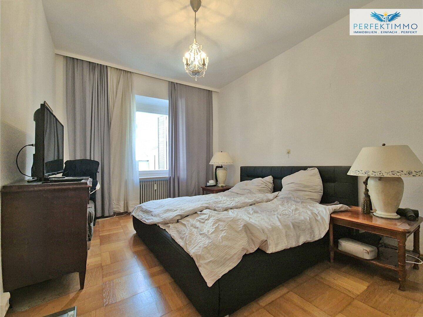 Helles Schlafzimmer mit Doppelbett, Parkettboden und Vorhängen am Fenster.