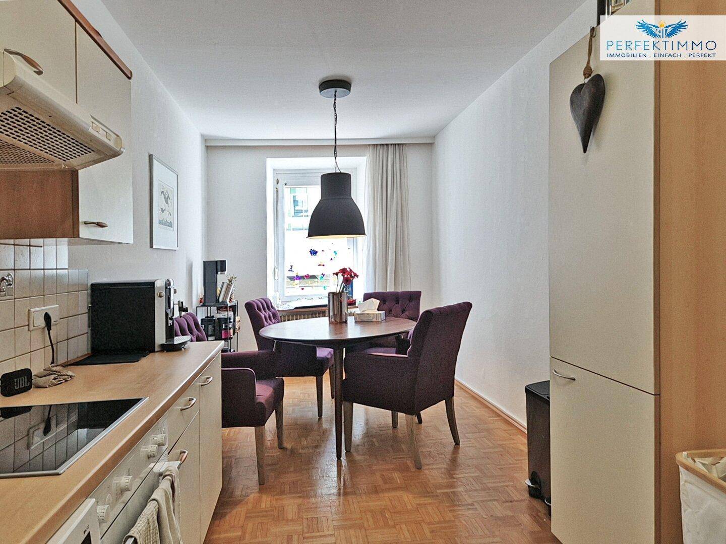 Essbereich in der Küche mit rundem Holztisch und lila Stühlen, beleuchtet von einer Pendelleuchte.