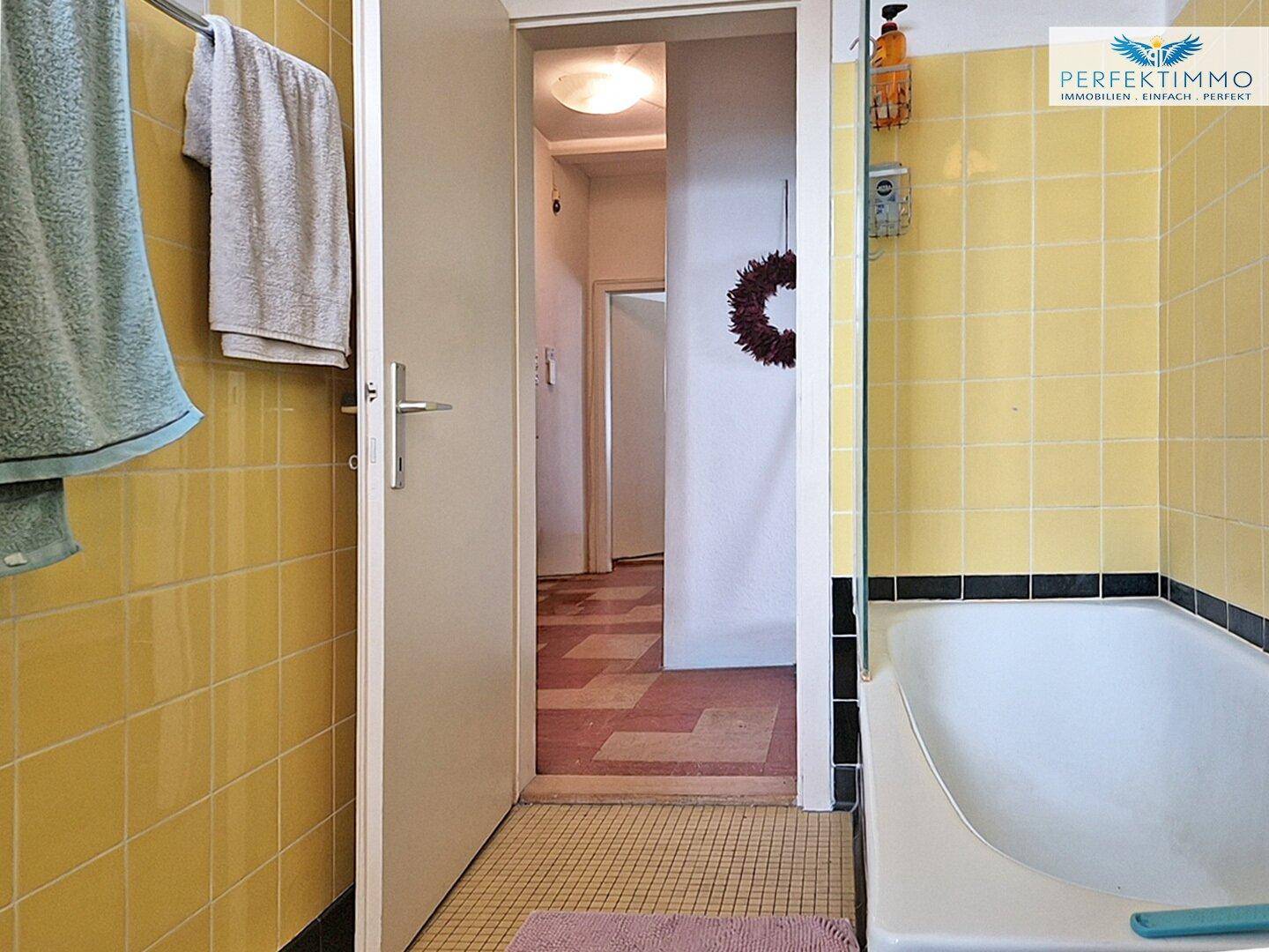 Badezimmer mit gelben Fliesen und Badewanne, Blick in den angrenzenden Flur.