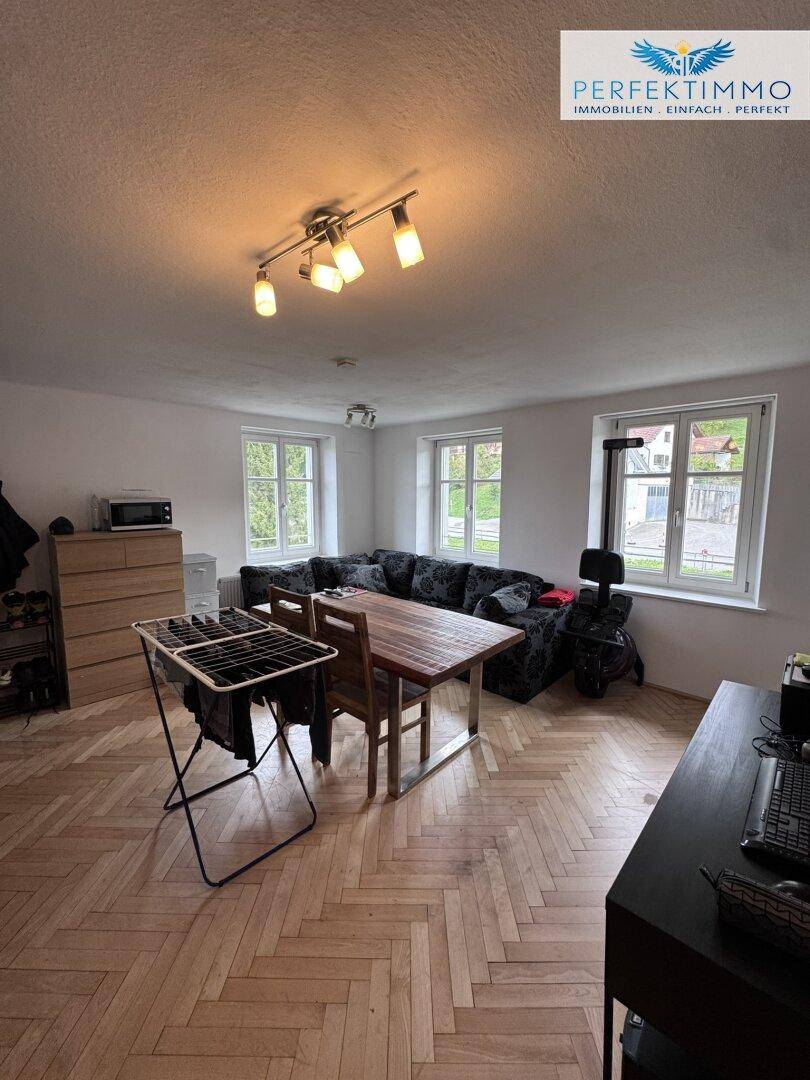 Heller Wohn- und Essbereich mit Fischgrätparkett, Sofa und Esstisch, gut beleuchtet durch Fenster.