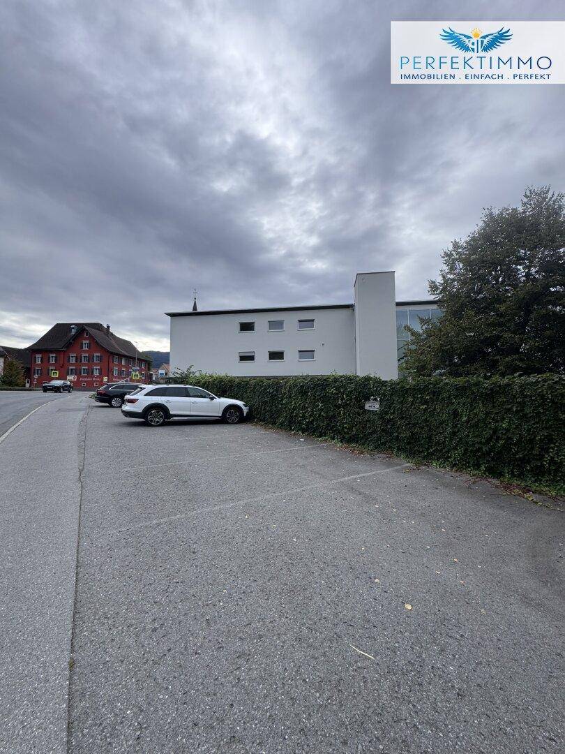 Außenbereich mit Parkplätzen und einer hohen Hecke, die das Grundstück von der Straße trennt.