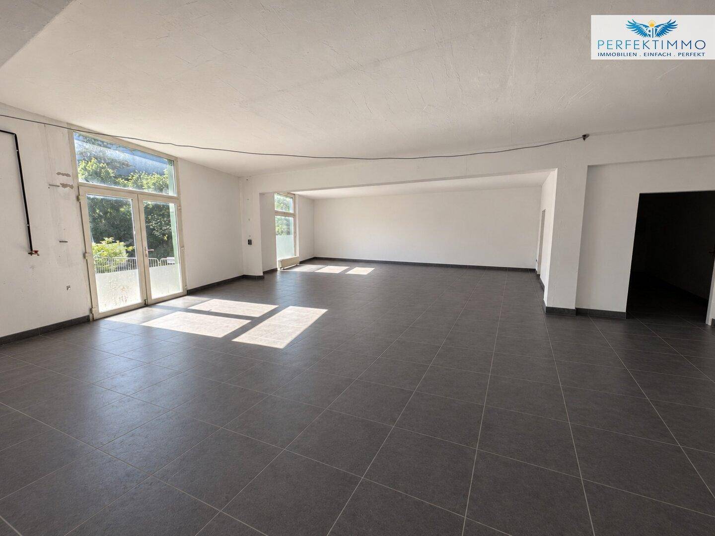 Weiterer Blick in den großen, leeren Gewerberaum mit viel Tageslicht und dunklen Fliesen.