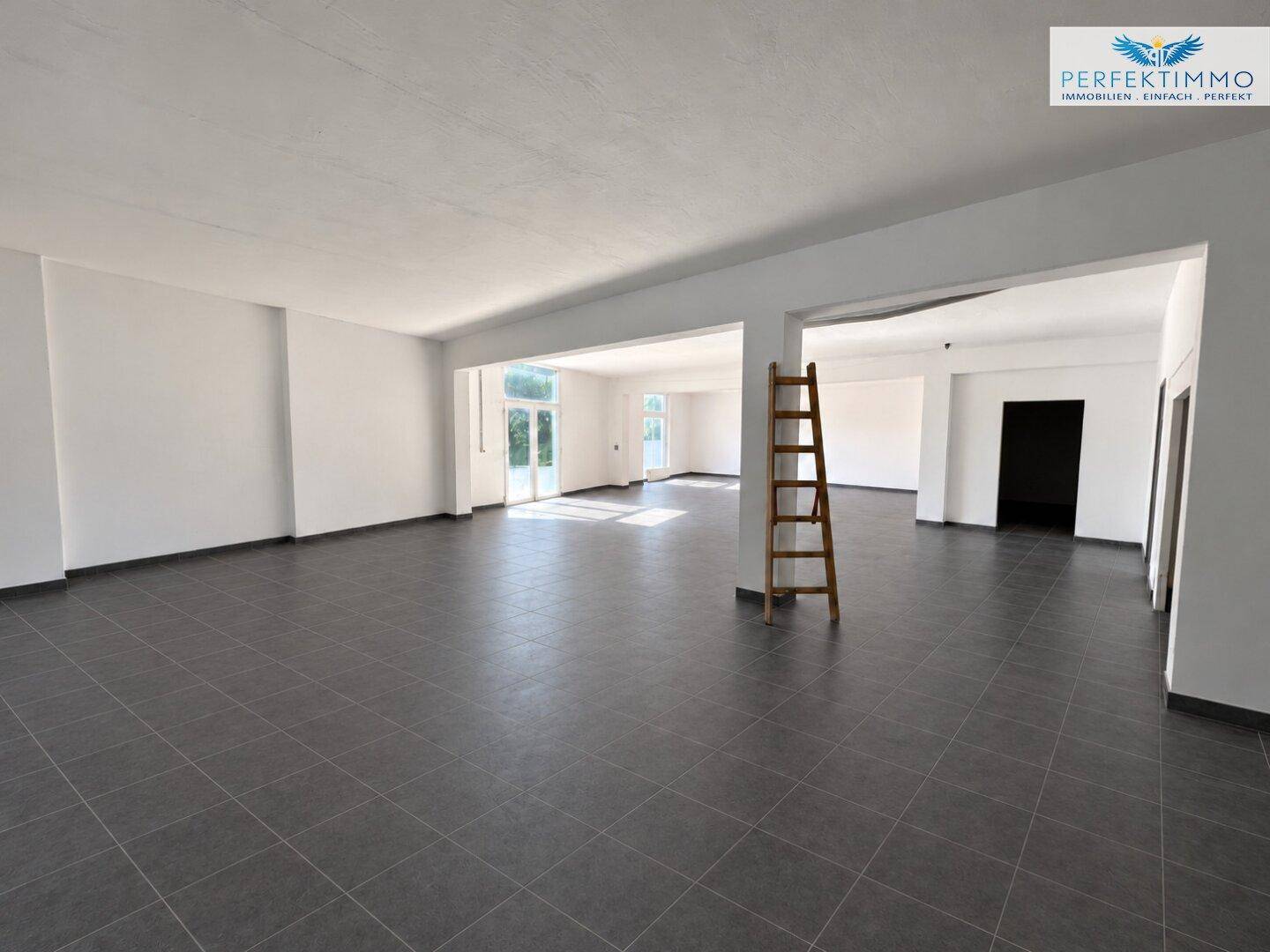 Offener Gewerberaum mit grauen Fliesen und einer Holzleiter, bereit für den Ausbau oder die Einrichtung.