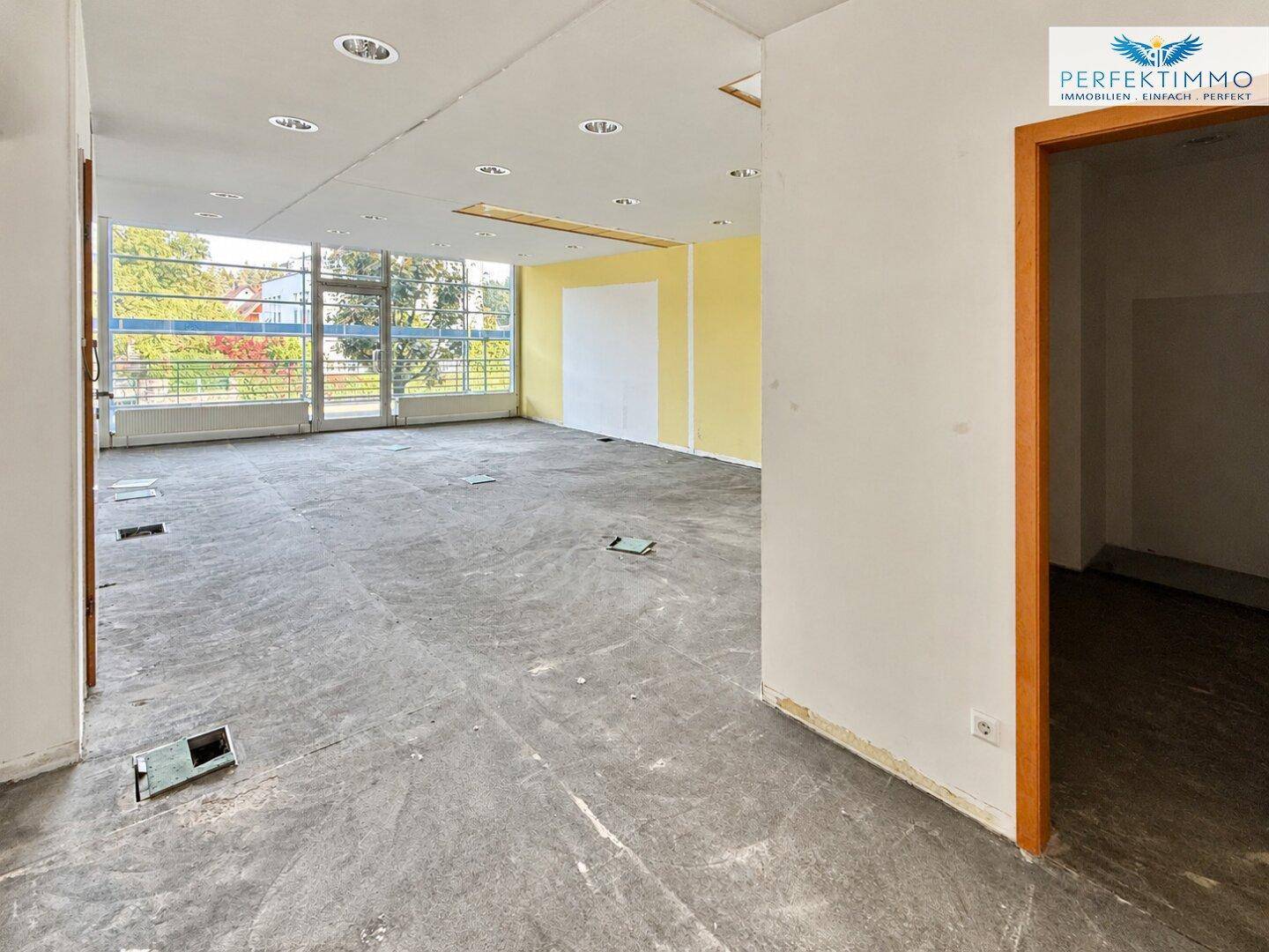 Großer, leerer Gewerberaum mit großen Fenstern und vorbereitetem Boden für neue Beläge, hell und offen.