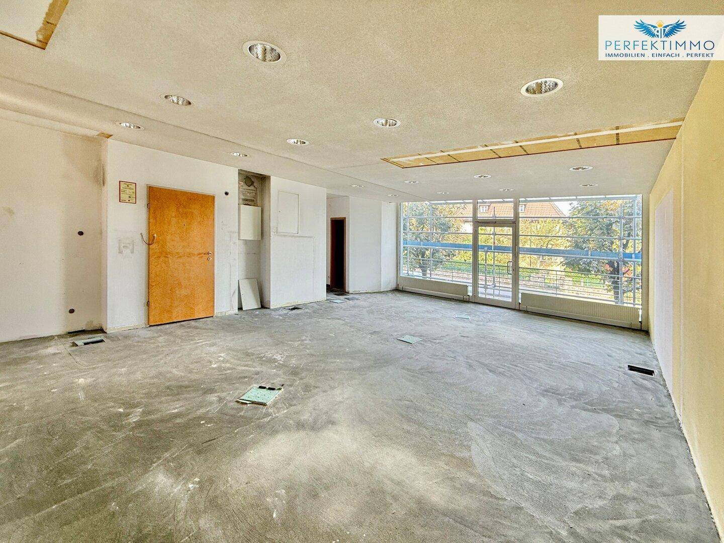 Weiter Blick in einen großen Gewerberaum mit vielen Fenstern und unfertigem Boden, ideal für Umbau.